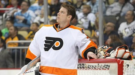 Mason leaning with left hand on the ice hockey post as he looks up to the right with no helmet on, in the white dominant Flyers dress.