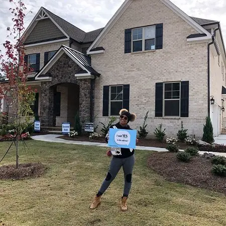 Eva Marcille posing on the lawn in front of her lovely new beige house holding a sign that said, "I said YES to the address!"