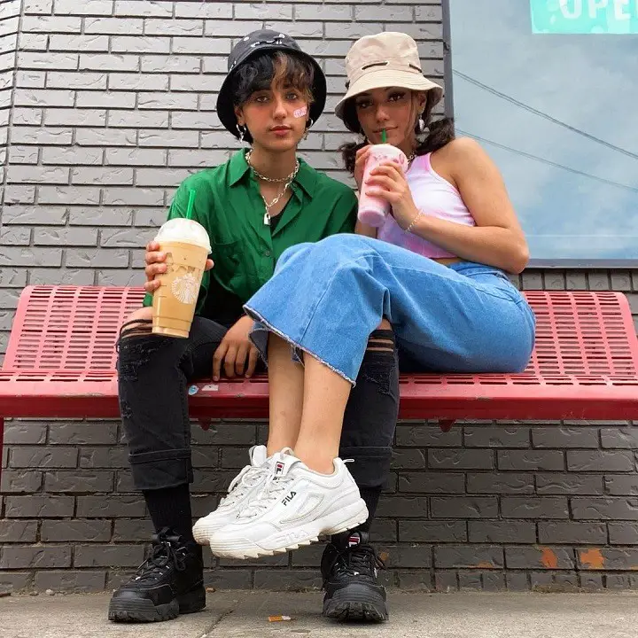 Noor Dabash (right) sipping a drink while crouching her legs on her sibling Diya Dabash's leg (left) while sitting on a red bench.