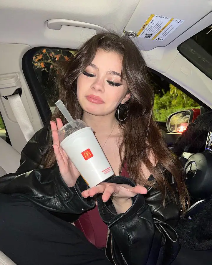 Addison Riecke presenting a McDonald drink, including its straw, inside a car while making a 'meh' face.