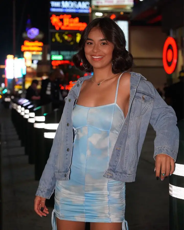 Giselle Lomelino in a blue/white dress leaning on a waist-level pole looking at the camera.