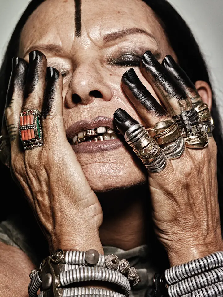 Michele Lamy posing for a photo for Interview Magazine showing her gold teeth with her hands on her cheeks and eyes closes.