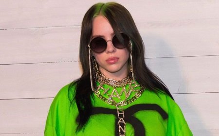 Billie Eilish Wearing a green top with jewelry in her ears and neck and round black spectacles.