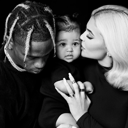 Kylie (right) kissing Stormi (center) on the cheek with Travis (left) looking down beside them in a B/W photo.