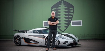 Christian von Koenigsegg in standing in front of his Agera RS folding arms and the brand logo behind him on a green background.