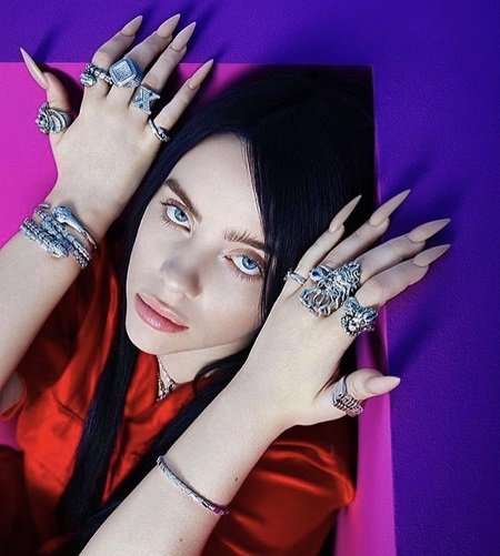 Billie lying on a bed (headshot) with her hands shown, every finger filled with rings. Her Net Worth is helping her.