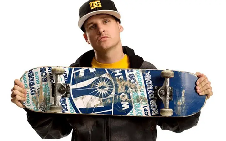 Rob Dyrdrek, who has a net worth of $50 million, holding a skateboard to his chest with the wheels shown and wearing a cap.