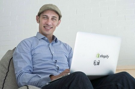 Tobi Lutke sitting on a couch with a laptop on his lap with the 'Shopify' sticker and smiling at the camera.