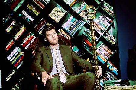 Colin Jost sitting on a chair with a mystical wand in his left hand and bookshelves behind him.