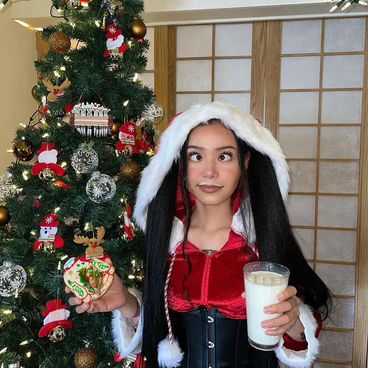 Bella Poarch in a Santa costume except the beard holding a glass of milk and making her signature face.