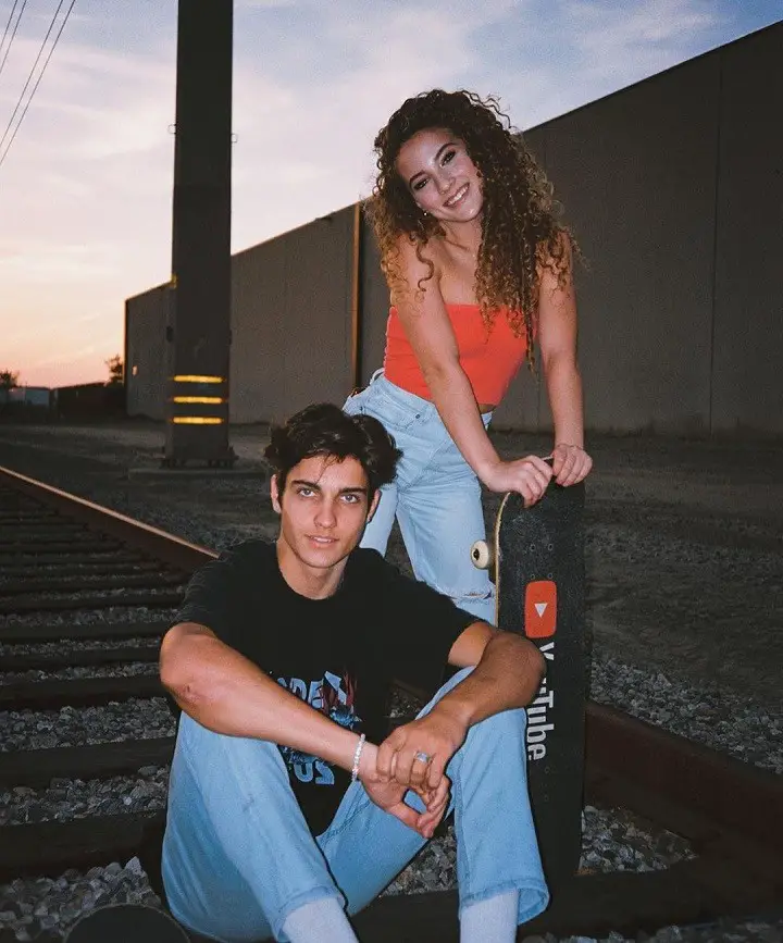 Dom Brack sitting on a railway track with Sofie Dossi standing behind him holding a skateboard with YouTube written on it.