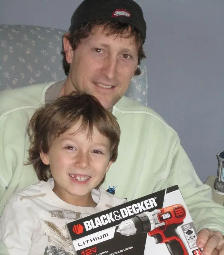 A young Skylar Gaertner sitting in front of his father with a Black + Decker drill case.