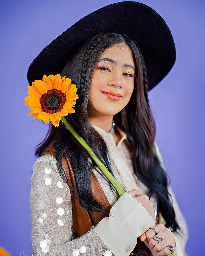 Niana Guerrero holding a sunflower in her right hand while wearing a hat over her braided hair.