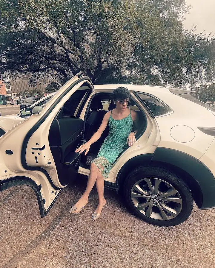 Nicholas Philmon wearing a green women's one-piece dress while posing as if he's getting out of a car wearing glasses.