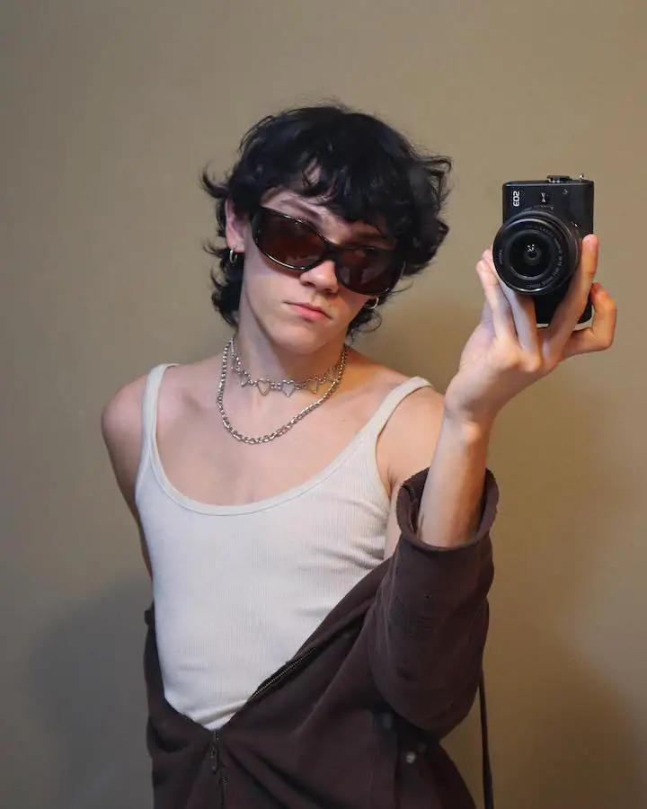Nicholas Philmon wearing glasses while holding a camera in his left hand wearing a white tank top.