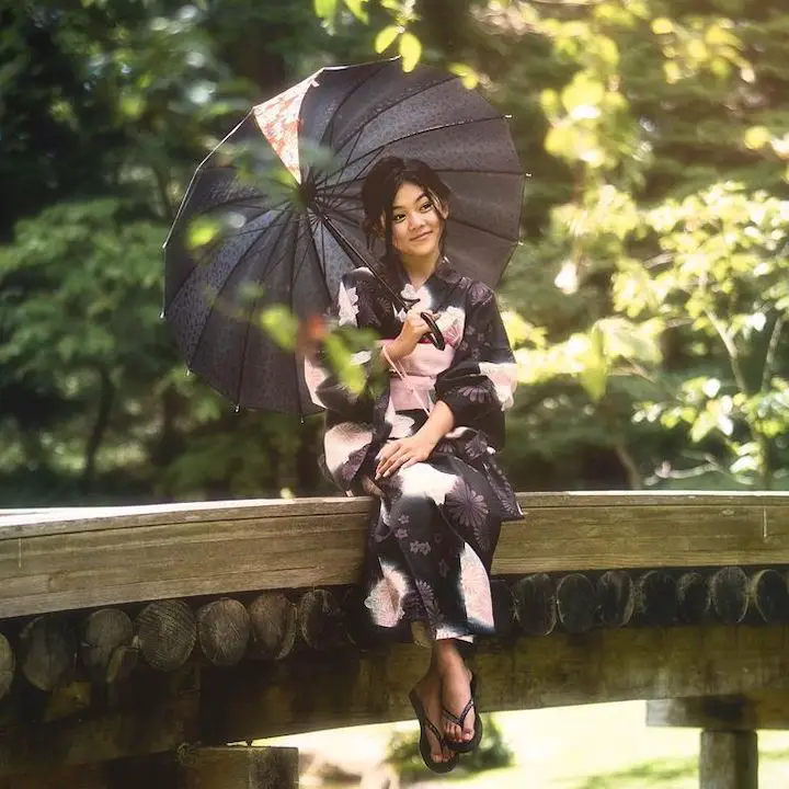 Momona Tamada sitting on a bridge railing in a traditional Japanese attire, Yukata, with an umbrella over her head.