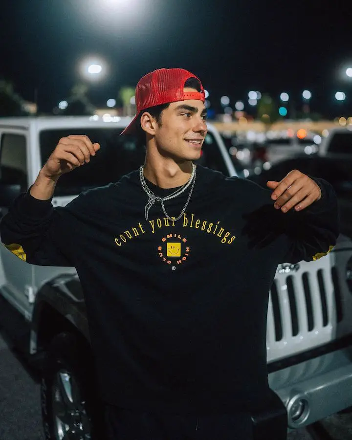 Jacob Rott of the Elevator Boys standing in front of a Jeep-like car looking to the side while wearing a red cap pn backwards and a loose-fit shirt that says 'Count Your Blessing' on it.
