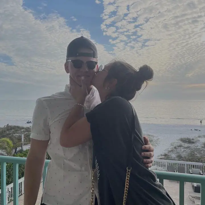 Signa Mae (right) kissing her boyfriend Carter Rinehart (left) on the cheek as he looks at the camera with the sea and the partly cloudy sky in the background.
