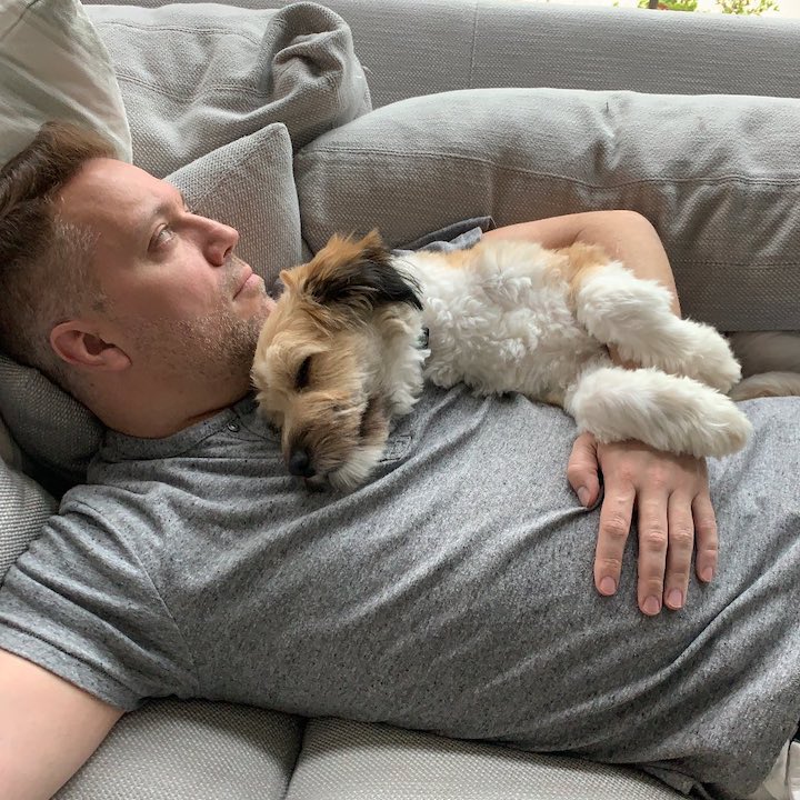 Shaun McKnight resting on a couch with their dog on top of him with its eyes closed.