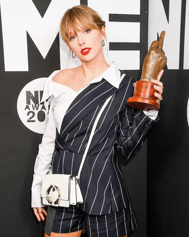 Taylor Swift holding an award that is a hand showing the finger, when showing her teeth-showing non-smile.