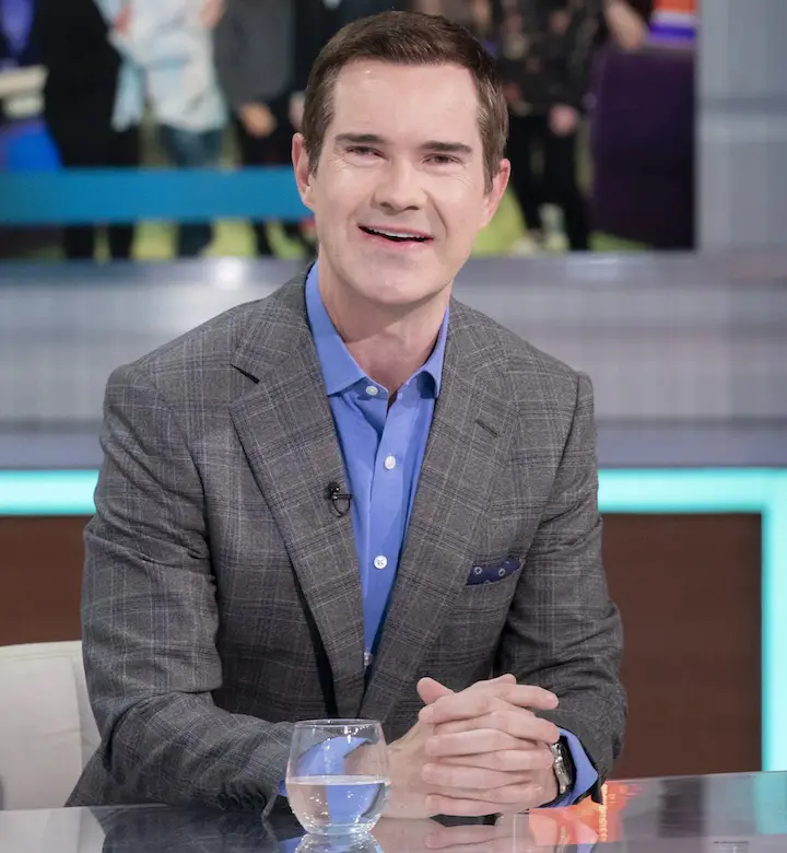 Jimmy Carr sitting beside a morning show desk in suit and a dapper smile on his face.