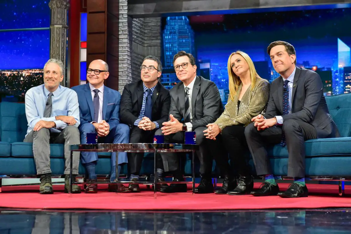 Stephen Colbert (third from right) sitting on an extended couch with five of his former colleagues from 'The Daily Show' including Jon Stewart (far left) and John Oliver (third from left).