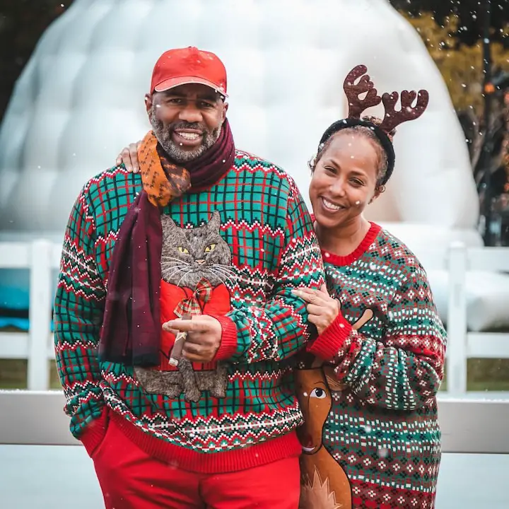 Steve Harvey (left) with his grey mustache-beard combo and his wife standing outside in Christmas-themed clothes smiling at the camera.
